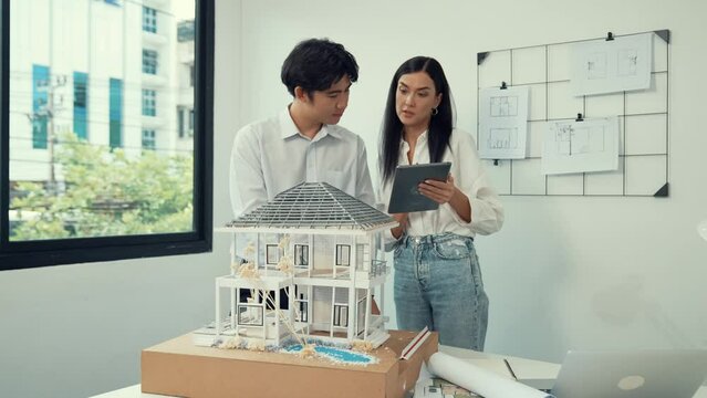 Beautiful young caucasian project manager brief asian engineer by using tablet while engineer measuring house model on meeting table with blueprint, architectural equipment scatter around. Immaculate