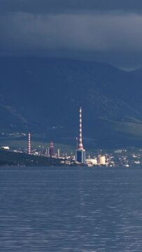 Oil and gas or petrochemical terminal on Krk island, Omisalj, Croatia. Towers with red and white stripes, a gas flare and wavy water against the Primorje-Gorski Kotar mountains on a cloudy afternoon.