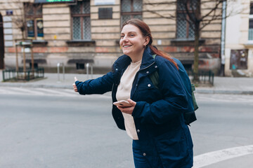 Stylish 30s woman with backpack holding smart phone and catching a taxi on the city street. Urban lifestyle concept.