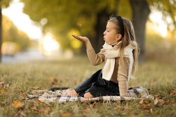 Stylish charming little child caucasian girl 5-6 years has fun outdoors, blows fallen leaves from her hand, sits on plaid on grass outdoors at autumn park. Children's day. Leisure activity on holiday