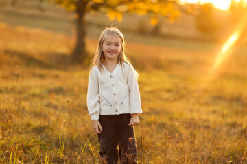 Children's day. Portrait of adorable little child caucasian girl 5-6 years smiles and looks at camera with crossed hands her hand outdoors in fall sunset lights. Leisure activity on autumn holiday.