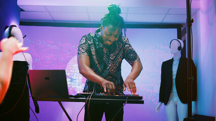 Young man mixing sounds on stage at party, having fun partying with group of friends at nightclub. Male DJ using audio station panel to mix electronic music under disco lights. Handheld shot.