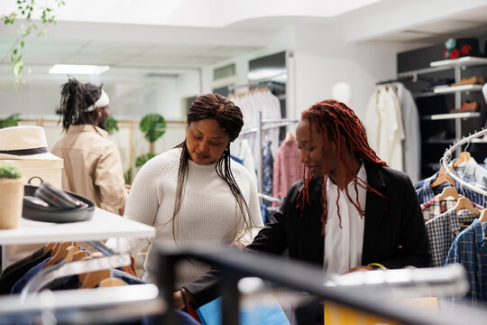 African american women friends browsing apparel and accessories in clothing store. Girlfriends checking garment for sale hanging on rack and shopping together in mall boutique - Powered by Adobe