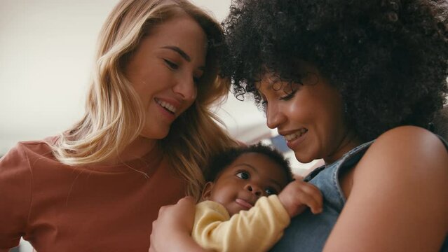 Same sex female couple or friends playing with baby sitting on sofa at home together - shot in slow motion