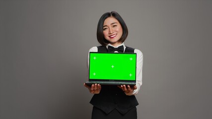 Receptionist showing greenscreen display on personal laptop, presenting isolated copyspace mockup on screen. Asian hotel employee holding pc with chromakey mockup template. Camera A.