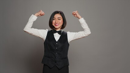 Asian hotel concierge flexing arms muscles in grey studio, showing off her strength and power in...