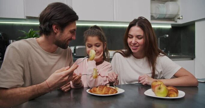 A happy brunette man with stubble in a beige T-shirt together with his brunette wife in a white T-shirt and a little girl daughter in pink clothes are having breakfast in a modern apartment at a black