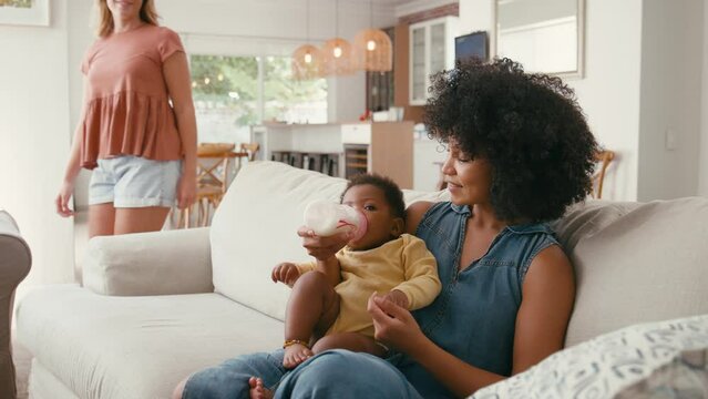 Same sex female couple or friends feeding baby sitting on sofa at home together - shot in slow motion