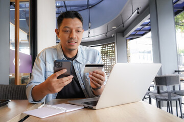 Asian man using laptop and mobile phone for shopping online with credit card in coffee shop online freelance business - obrazy, fototapety, plakaty