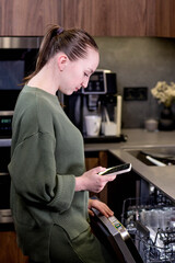 Woman is holding a phone with the smart home program running, remotely controlling the dishwasher. The concept of a smart kitchen at home