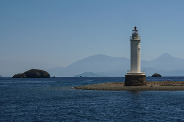 A lighthouse on a small island, the background features a clear blue sky and mountains, clear water