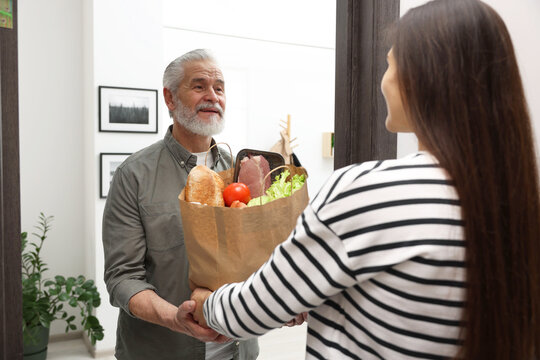 Courier giving paper bag with food products to senior man indoors