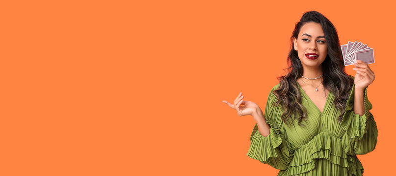 Rich young woman with poker playing cards showing something on orange background with space for text