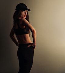 Sport sexy body beautiful slim woman with long hair posing in black sport bra, leggings, summer cap showing the shoulders, arms, standing on studio background with empty copy space. Healthy lifestyle