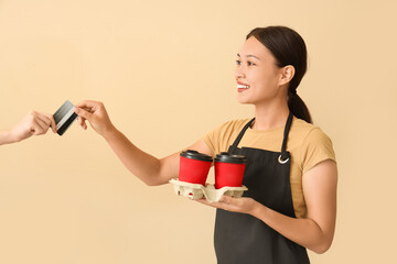 Female Asian barista with cups of coffee taking credit card on beige background