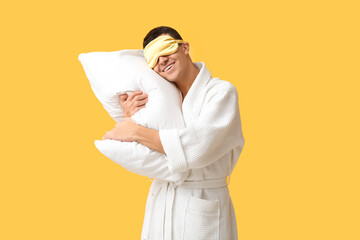 Portrait of handsome young man in bathrobe with sleeping mask and pillow on yellow background
