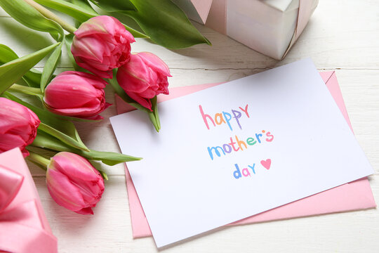 Envelope with festive postcard, gift boxes and tulips on white wooden background. Happy Mother's Day