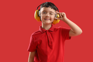 Cute little builder in safety goggles and hearing protectors winking on red background