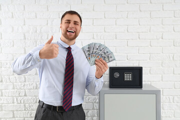 Happy young man with safe boxes and money showing thumb-up gesture on white brick background