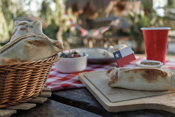 Chilean pino empanadas on wooden board and table in park, with pebre bowl, wicker basket, Chilean flag, glass and red and white checkered kitchen towels. Typical Chilean food
