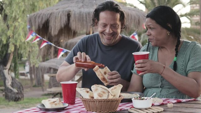 A couple shares an intimate outdoor meal in the park. Both are happy and laughing, about to bite into Chilean pino empanadas. Chilean flag, Independence Day celebrations