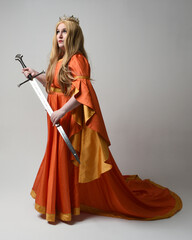 Full length portrait of plus sized woman blonde hair, wearing historical medieval fantasy gown, golden crown of royal queen. Standing pose holding sword weapon, isolated studio background.