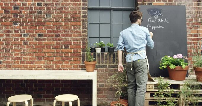 Board, writing and young barista at coffee shop with creative and art menu list. Startup, small business and back of male waiter creating chalk sign for special outside the restaurant, cafe or diner.