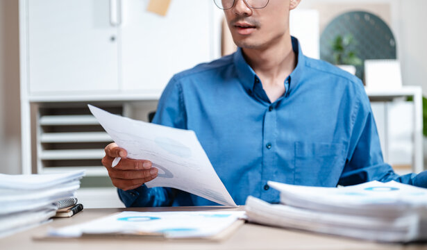 contract, document, executive, job, manager, occupation, paperwork, report, working, information. A man is sitting at a desk with a pile of papers in front of him. He is reading a piece of paper.