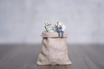Closeup image of miniature businessman sitting on a large sack overflowing with U.S. dollar bills....