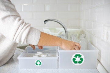 In the kitchen, a woman rinses plastic bottles and sorts them into a box with recycling signs. A...