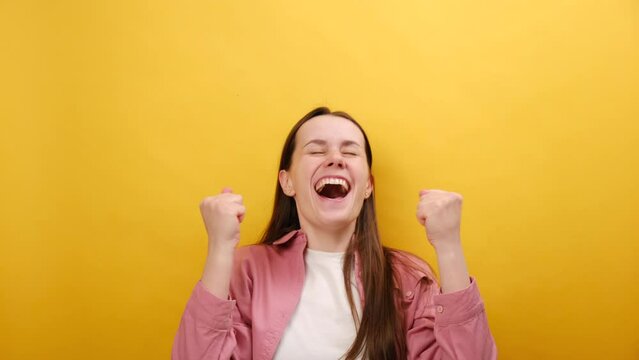 Portrait of emotional young woman watches lottery draw or sports game. Lucky female shouts happily and waves hands rejoicing in victory of favourite team, posing isolated over yellow background wall