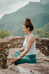 Naklejka premium A Woman Achieves Inner Peace through Yoga and Meditation in Lotus Seat, Surrounded by the Tranquil Ambiance of an Old Stone Wall amidst the Lush Greenery of a Forest Backdrop