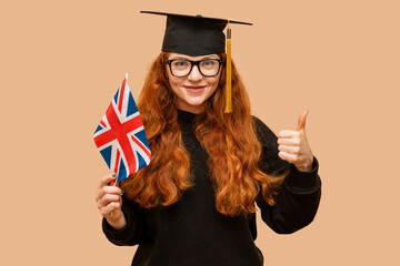 Female student wearing UK flag glasses showing thumbs up, wearing bachelor's cap, standing on pink...