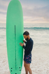 Young  bearded man with surfboard standing near a beach. Man with surfing board outdoors on a summer day.