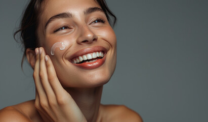 A beautiful woman with radiant skin, smiling and happy while gently applying cream to her face on a gray background