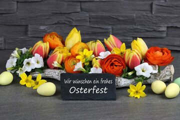 Blumen mit Ostereiern und einer Schiefertafel mit dem Schriftzug Frohe Ostern.	