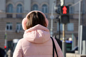 Girl with pink headphones standing on red traffic light background. Headset, listening to music and city life concept