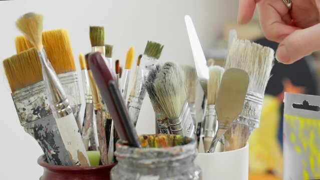 Artist checking paintbrushes and palette knifes in glass before drawing picture. Unrecognizable young artist choosing brushes. Artwork. Tools for painting. Close-up in 4K, UHD