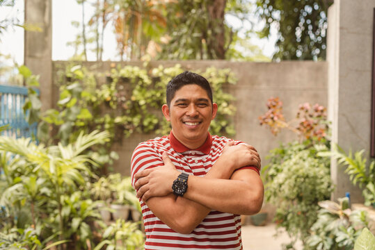 Content Asian man in striped shirt hugging himself in a sunny garden