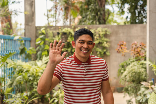 Cheerful Asian man waving hello in a lush outdoor setting, exuding friendliness and warmth