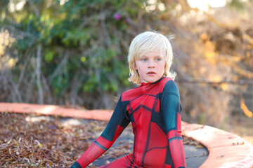 Boy wearing red superhero suit sitting on trampoline covered in autumn leaves