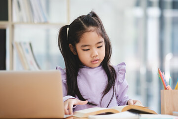 A young girl is sitting at a desk with a laptop and a book