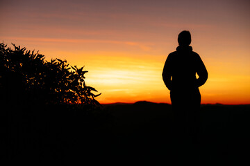 A silhouetted female stands with a vibrant coloured sunrise in front of her.