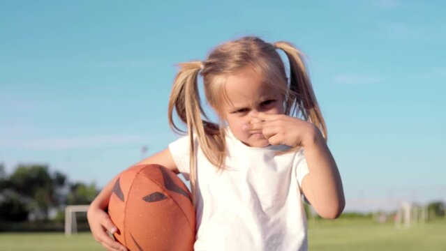 A little girl holding a rugby ball and making a hand gesture

