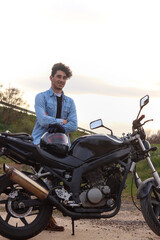 Fototapeta na wymiar Young Caucasian man with curly hair standing next to the motorcycle with crossed arms looking forward, vertical image. Lifestyle concept