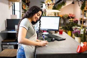 A young Latina woman works inside a pretty flower shop dressed in an apron.The woman is writing in...