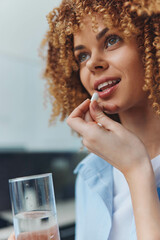 Curly haired woman taking a pill with a glass of water in hand, health and wellness concept