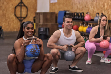 Group of multiracial smiling people doing squats with kettlebell dumbbells, in a row, inside a gym, sport dressed. Fitness concept