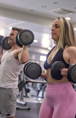 Fototapeta na wymiar Diverse couple smiling multiracial exercising their arms with dumbbells in a gym, vertical image. Fitness concept with normal people