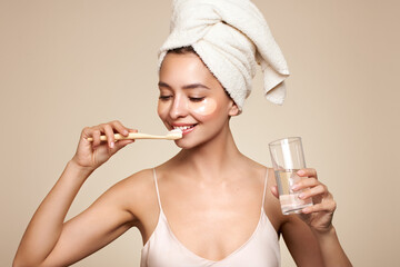 Image of a young happy positive woman in towel and with cosmetic patches on the eyes, brushing her...